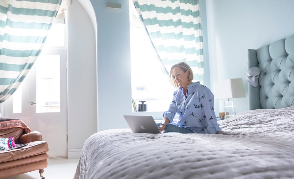 nainen istuu sängyllä makuuhuoneessa läppärin kanssa